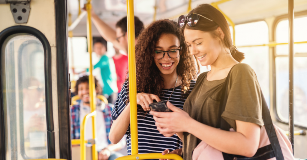 Two young women smiling and looking at a smartphone while riding a bus, with other passengers visible in the background.