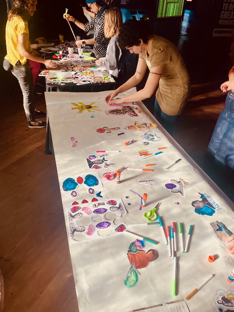 A group of people engaged in an art activity, creating colorful drawings and collages on a long table covered in paper with various markers, stickers, and crafts.