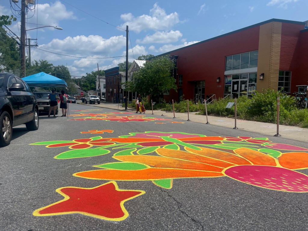 Colorful floral mural painted on a street, with vibrant flowers and leaves in shades of orange, yellow, green, pink, and red. People are visible in the background, along with a blue tent and parked cars.
