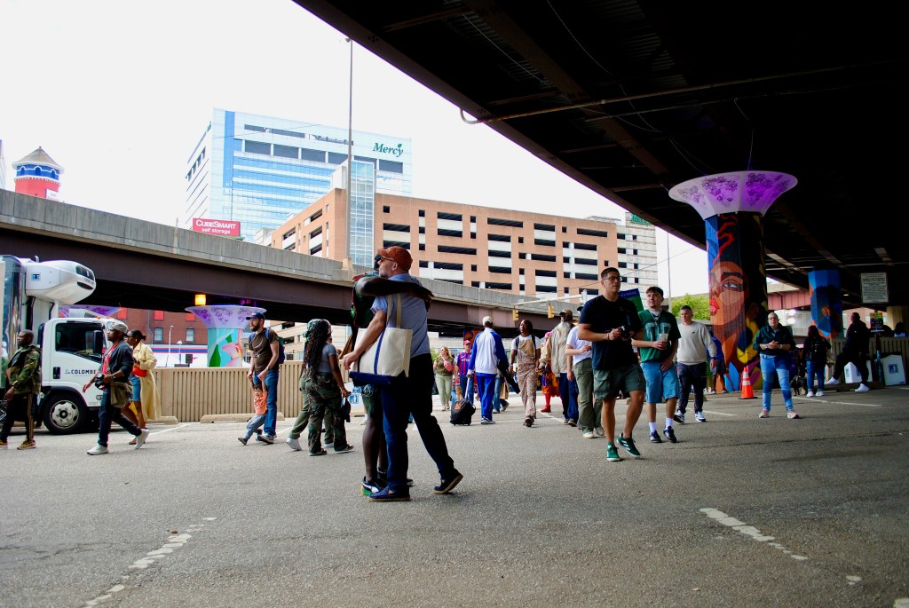 A diverse group of people walking under an overpass with colorful murals and tall buildings in the background. In the foreground, two people warmly greet each other with a hug.